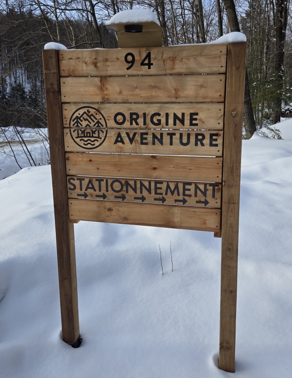 Snow covered entrance sign for Origine Aventure with parking directions.